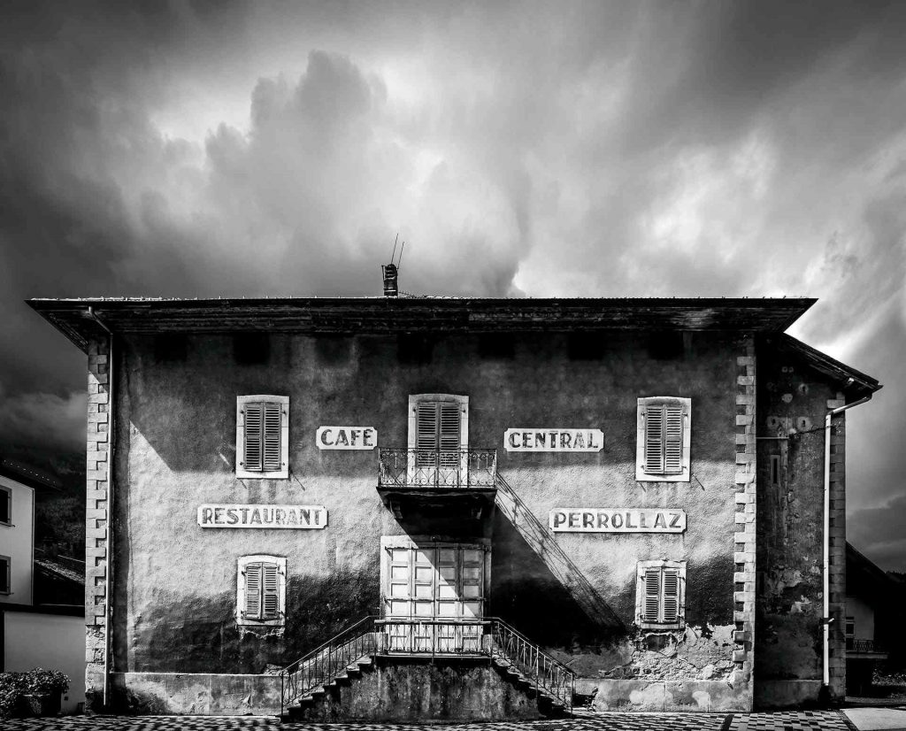 Ce que les pierres nous murmurent © Norbert Bardin Photo en noir et blanc de Norbert Bardin de la série "Ce que les pierres nous murmurent" de l'extérieur de la Maison forte de Loche à Magland. La bâtisse moyenâgeuse occupe le tiers inférieur de la photo et joue sur une symétrie quasi-parfaite cassée par les nuages aux lignes courbes du ciel.