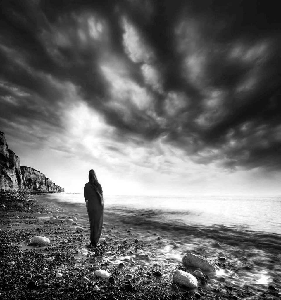 Née du vent, de la mer et du granit © Norbert Bardin Photo en noir et blanc de Norbert Bardin de la série "Dialogue du crétacé" prise sur les plages d'Etretat de la silhouette d'une femme debout sur les galets proches de la mer avec les falaises sur sa gauche sous un ciel gris moutonneux s'éclaircissant vers l'horizon.