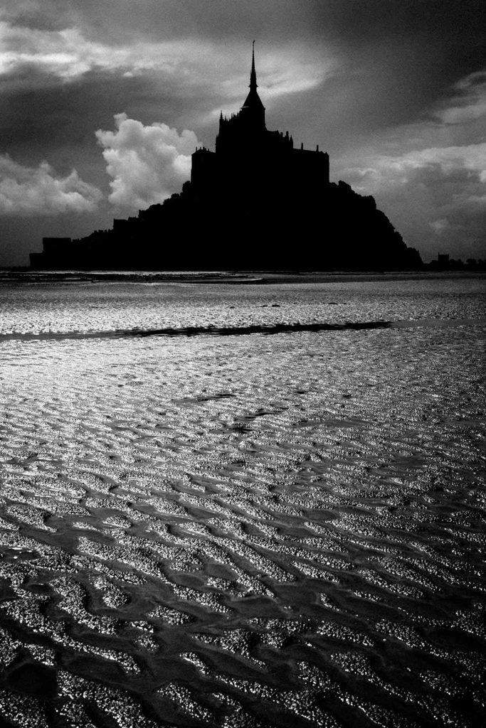 Empreintes marines © Norbert Bardin Photo en noir et blanc de Norbert Bardin de la série "Empreintes marines" du Mont-Saint-Michel dont la silhouette noire se détache sur un ciel nuageux duquel transperce des rayons de soleil. La lumière joue sur le sable et le peu d'eau laissé par la mer qui s'est retirée.