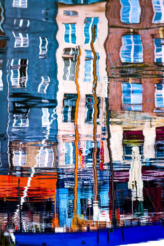 L'imagier de l'eau © Norbert Bardin Photo en couleur de Norbert Bardin de la série "L'imagier de l'eau" de façades de maison qui se reflètent dans la mer. Au premier plan un bateau à la coque bleue se distingue clairement. Les ondulations assez horizontales brouillent les lignes des éléments des maisons et du bateau.